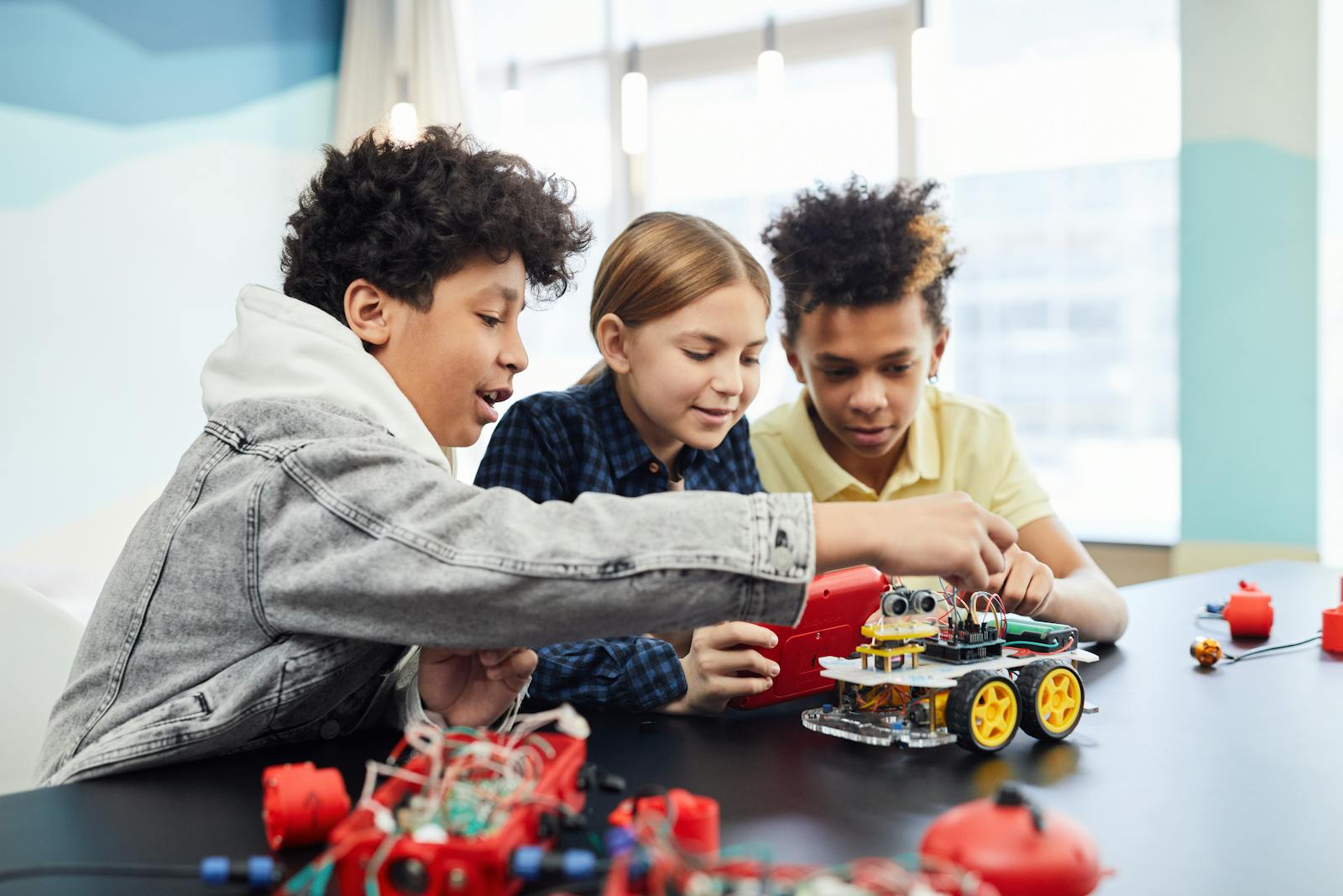 Trys mokiniai kartu konstruoja roboto modelį prie klasės stalo.